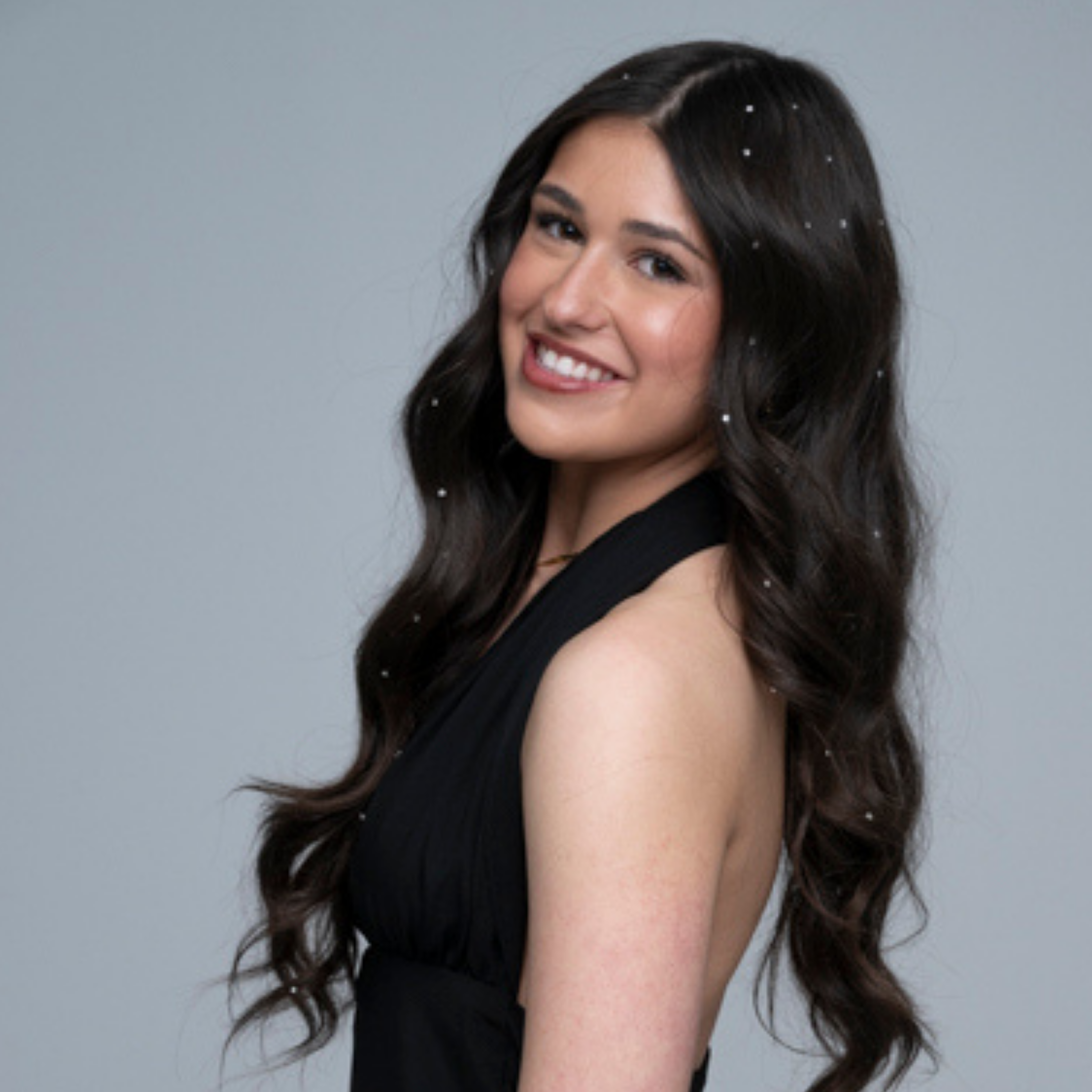 Teen with long, wavy hair wearing a black dress and blinger gems in her hair against a gray background